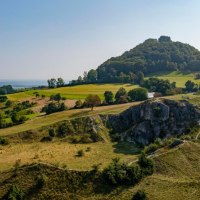 Grüne Hügel und Bäume unter klarem Himmel, mit einem markanten Felsen im Vordergrund. Im Hintergrund ist ein bewaldeter Hügel zu sehen., © TMBW