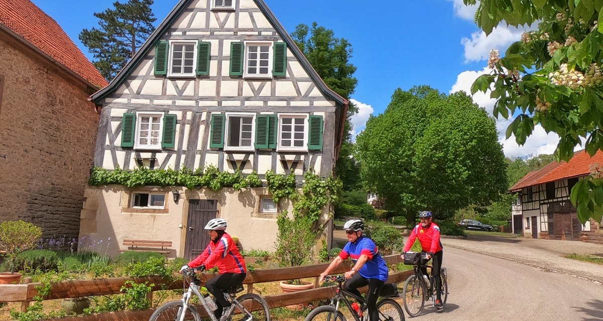 Drei Radfahrer in bunter Kleidung fahren an einem malerischen Fachwerkhaus vorbei. Der Himmel ist blau mit weißen Wolken., © Land der 1000 Hügel - Kraichgau-Stromberg Drei Radfahrer in bunter Kleidung fahren an einem malerischen Fachwerkhaus vorbei. Der Himmel ist blau mit weißen Wolken., © Land der 1000 Hügel - Kraichgau-Stromberg