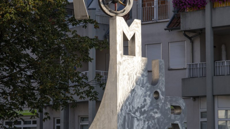Der Katz- und Mausbrunnen in Bottwartal zeigt eine moderne Skulptur mit Buchstaben und geometrischen Formen vor einem Gebäude mit roten Dachziegeln., © Stuttgart Marketing Der Katz- und Mausbrunnen in Bottwartal zeigt eine moderne Skulptur mit Buchstaben und geometrischen Formen vor einem Gebäude mit roten Dachziegeln., © Stuttgart Marketing