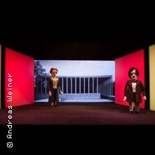 Zwei Puppen auf einer B&uuml;hne mit architektonischem Hintergrund. Die Beleuchtung ist in Rot- und Gelbt&ouml;nen gehalten., &copy; links im Bild