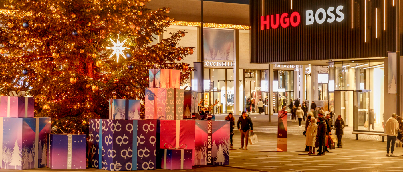 Weihnachtlich geschmückter Platz mit großem Baum und Geschenken vor dem Hugo Boss Geschäft. Menschen flanieren in der festlichen Atmosphäre., © OUTLETCITY AG