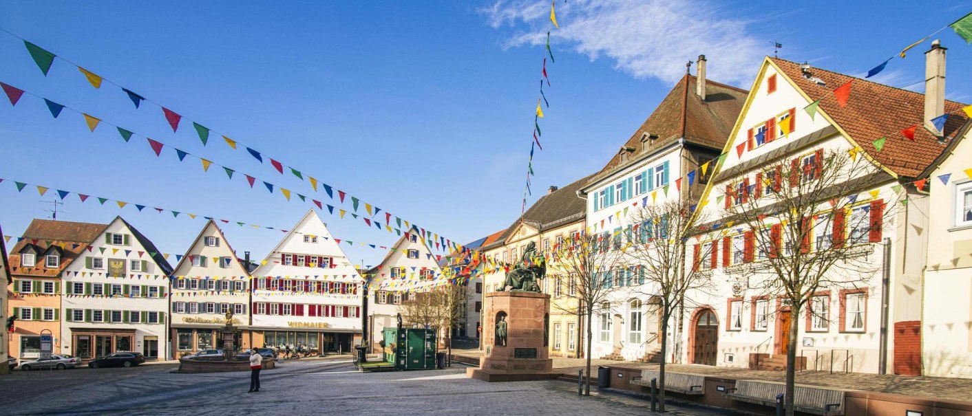 Der Marktplatz von Weil der Stadt zeigt historische Gebäude und bunte Wimpel unter blauem Himmel. Eine Statue steht zentral auf dem Platz., © SMG, Sarah Schmid Der Marktplatz von Weil der Stadt zeigt historische Gebäude und bunte Wimpel unter blauem Himmel. Eine Statue steht zentral auf dem Platz., © SMG, Sarah Schmid