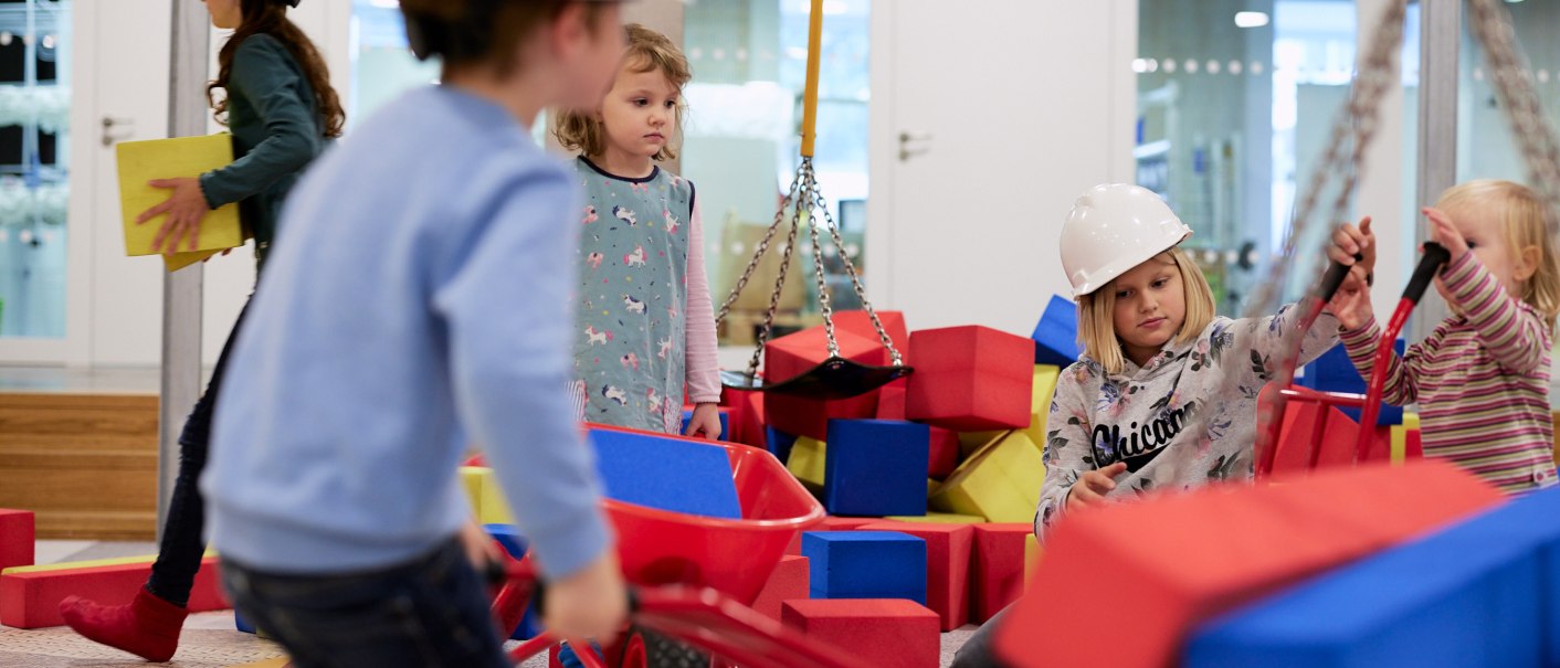 Kinder mit Bauhelmen spielen auf einer Indoor-Baustelle mit bunten Bauklötzen und Schubkarren., © Julia Ochs Kinder mit Bauhelmen spielen auf einer Indoor-Baustelle mit bunten Bauklötzen und Schubkarren., © Julia Ochs