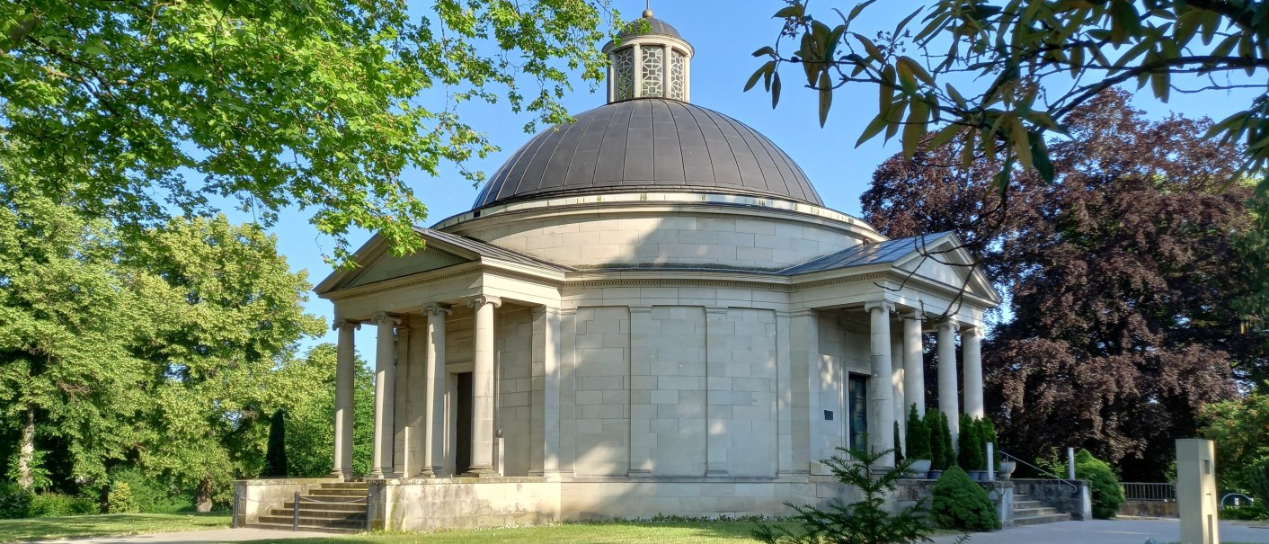 Frasch Mausoleum auf dem Friedhof in Gaildorf: helles Steingeb&auml;ude mit grauer Kuppel umgeben von B&auml;umen und Gr&uuml;nfl&auml;chen. Von S&auml;ulen getragene Vord&auml;cher sch&uuml;tzen zwei Eingangst&uuml;ren, zu denen Stufen hinauff&uuml;hren., &copy; Petra Natzkowski