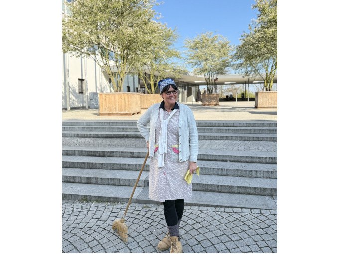 Frau mit Kopftuch und Besen steht lächelnd auf einem gepflasterten Platz vor modernen Gebäuden und Bäumen., © WTM GmbH Waiblingen Frau mit Kopftuch und Besen steht lächelnd auf einem gepflasterten Platz vor modernen Gebäuden und Bäumen., © WTM GmbH Waiblingen