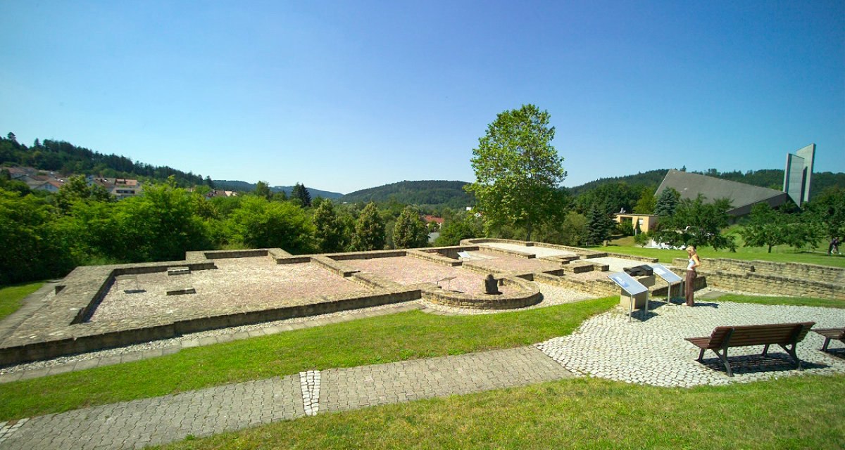 Ruinen des Kastellbads Schirenhof mit Informationstafeln, einer Person und einer modernen Kirche im Hintergrund. Gr&uuml;ne Landschaft und blauer Himmel., &copy; Stadtverwaltung Schw&auml;bisch Gm&uuml;nd