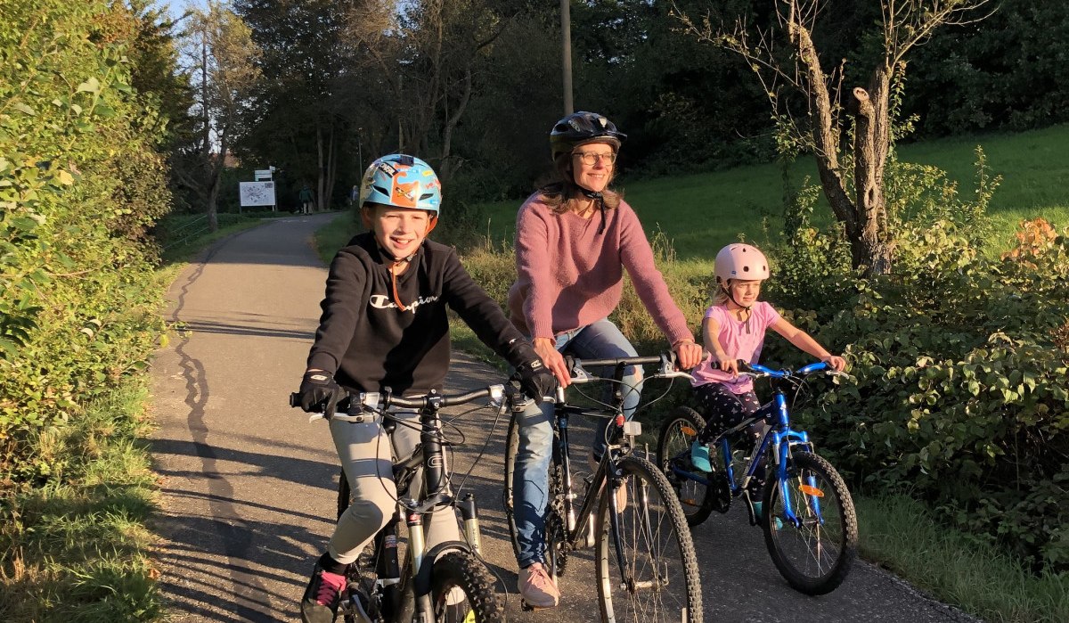 Drei Personen auf Fahrrädern fahren bei sonnigem Wetter auf einem Radweg durch eine grüne Landschaft., © T&M