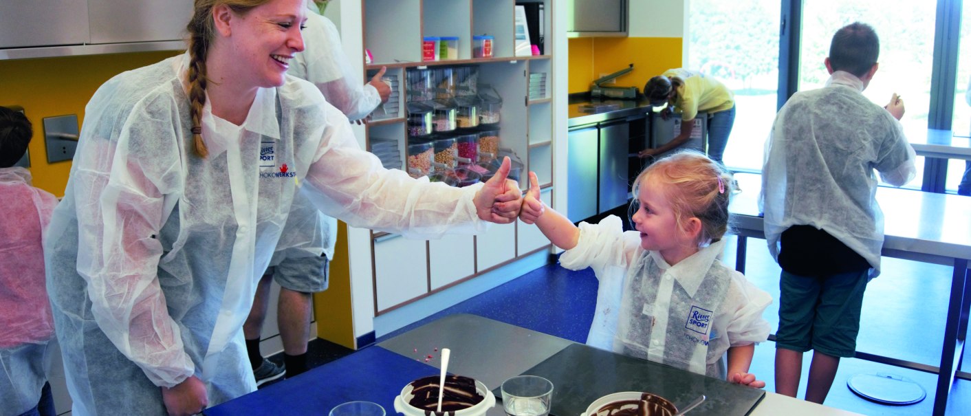 Erwachsene und Kind in Schutzkleidung geben sich in einer Schokoladenwerkstatt l&auml;chelnd die Daumen hoch. Sch&uuml;sseln mit Schokolade stehen auf dem Tisch., &copy; Alfred Ritter GmbH & Co. KG