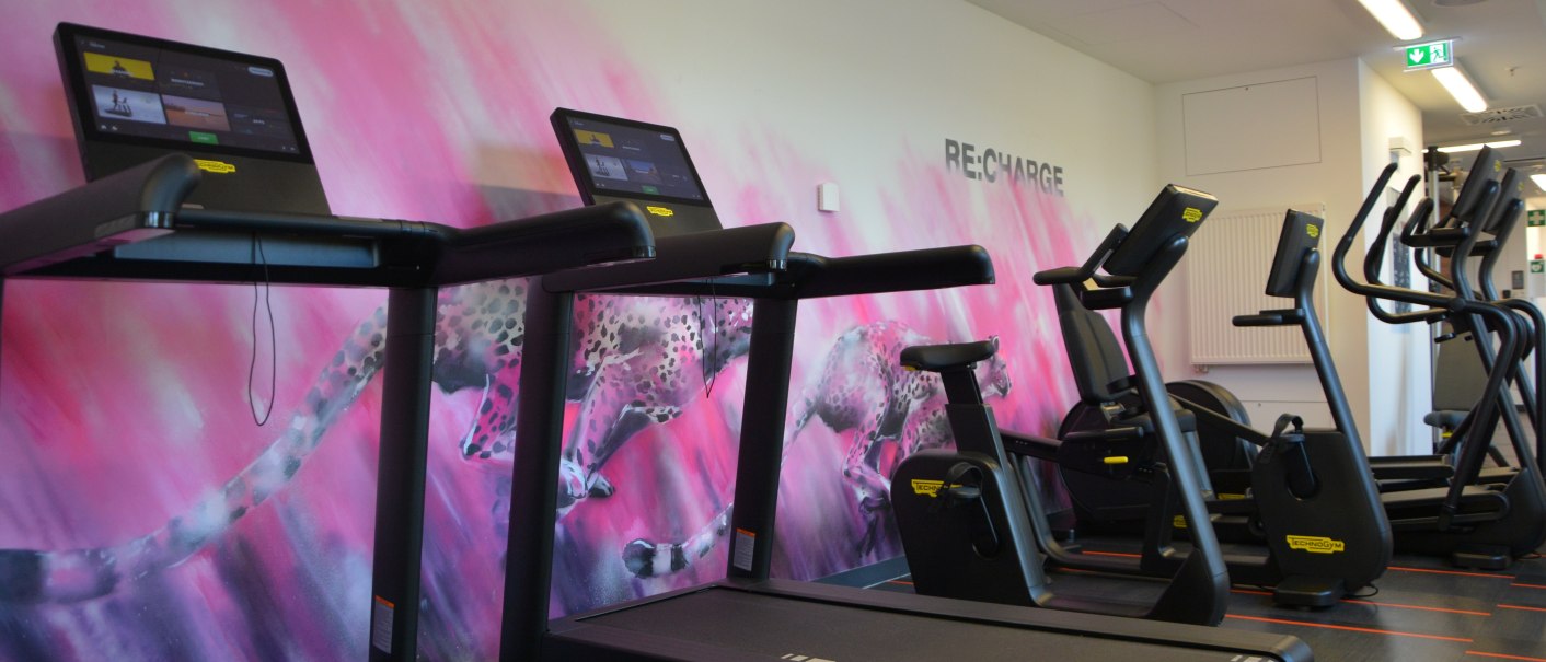 Ein Fitnessstudio mit modernen Laufb&auml;ndern und Crosstrainern vor einer rosa Wand mit einem Gepardenmotiv. Der Raum wirkt einladend und motivierend., &copy; Aloft Stuttgart