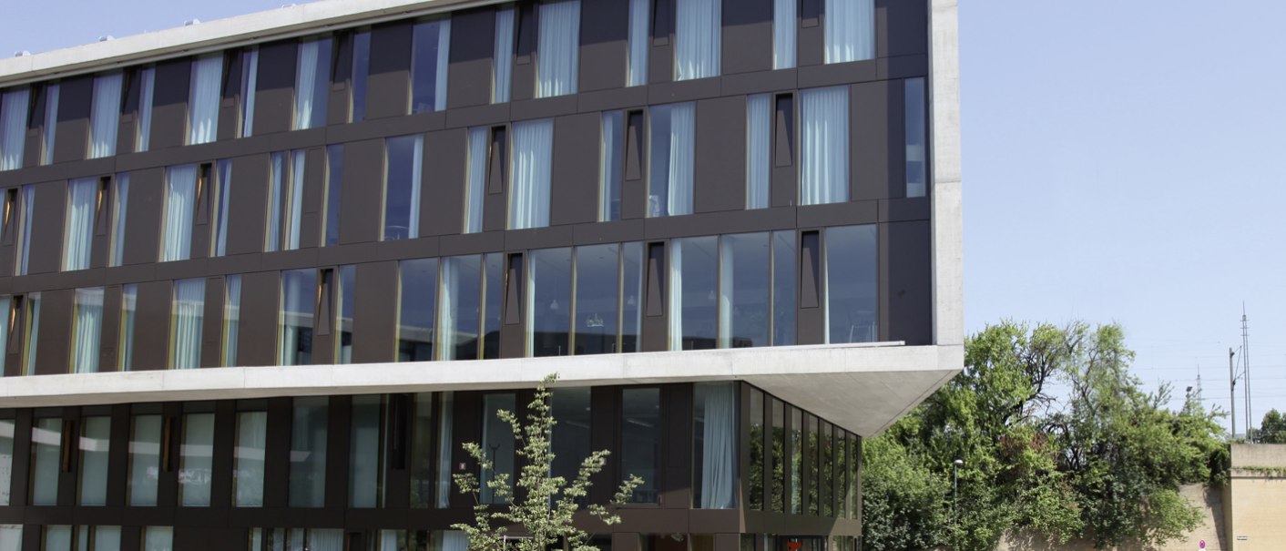 Modernes Gebäude mit Glasfassade, Terrasse und Bäumen im Vordergrund. Jugendherberge Stuttgart Neckarpark bei klarem Himmel., © DJH Landesverband Baden-Württemberg e.V.