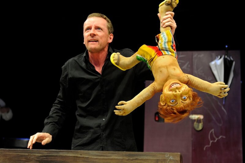 Ein Mann in schwarzem Hemd hält eine bunte Puppe kopfüber auf einer Bühne. Im Hintergrund sind Requisiten sichtbar., © Stadt Winnenden