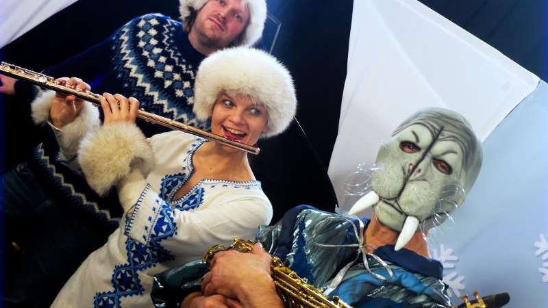 Drei Personen in Winterkleidung spielen Flöte und Saxophon, eine trägt eine Tiermaske. Hintergrund mit Schneeflocken und weißen Dreiecken., © Backnang Drei Personen in Winterkleidung spielen Flöte und Saxophon, eine trägt eine Tiermaske. Hintergrund mit Schneeflocken und weißen Dreiecken., © Backnang
