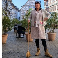 Eine Frau in traditioneller Kleidung mit Besen steht auf einem gepflasterten Platz vor Fachwerkhäusern. Sie trägt eine Mütze und Sonnenbrille., © WTM GmbH Waiblingen