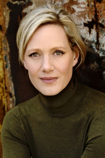 Eine Frau mit blondem Haar und gr&uuml;nem Rollkragenpullover blickt direkt in die Kamera. Der Hintergrund ist rustikal und texturiert.