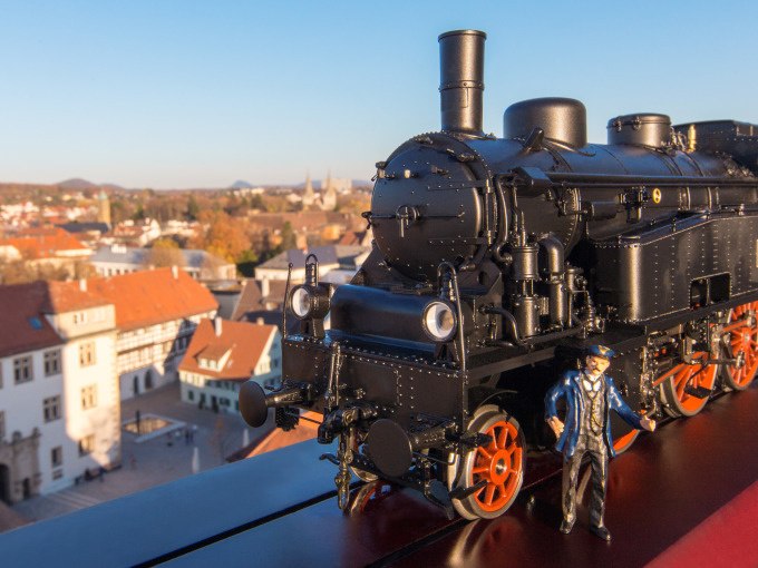Eine schwarze Modelllokomotive und eine Lokf&uuml;hrerfigur stehen auf einem Gel&auml;nder. Im Hintergrund ist eine Stadtlandschaft bei klarem Himmel zu sehen., &copy; Stadt G&ouml;ppingen