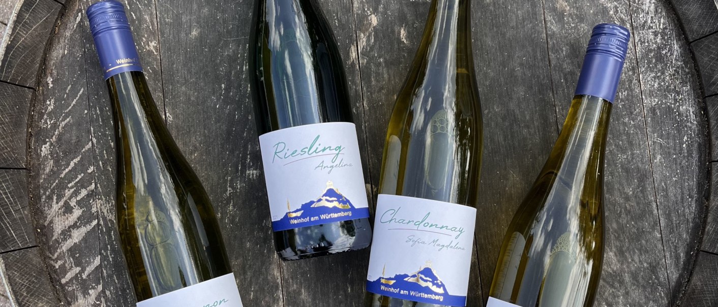 Vier Weinflaschen liegen auf einem Holzfass. Die Etiketten zeigen "Riesling" und "Chardonnay" vom Weinhof am Württemberg., © Weinhof Zaiß