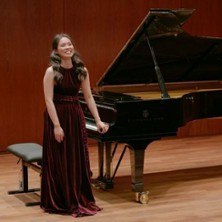 Eine Frau in einem eleganten, weinroten Kleid steht l&auml;chelnd neben einem Fl&uuml;gel auf einer B&uuml;hne. Der Raum hat eine warme, h&ouml;lzerne Atmosph&auml;re., &copy; links im Bild