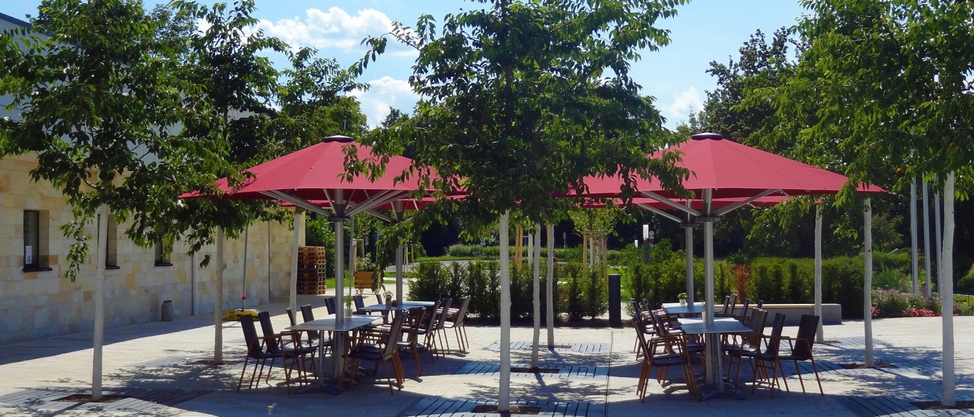 Außenterrasse des Restaurants Friedrichs in Beuren mit roten Sonnenschirmen, Tischen und Stühlen, umgeben von Bäumen und Pflanzen., © Tourist-Info Beuren
