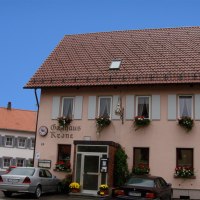 Gasthaus Krone mit blumengeschmückten Fenstern, zwei geparkte Autos davor, blauer Himmel im Hintergrund.