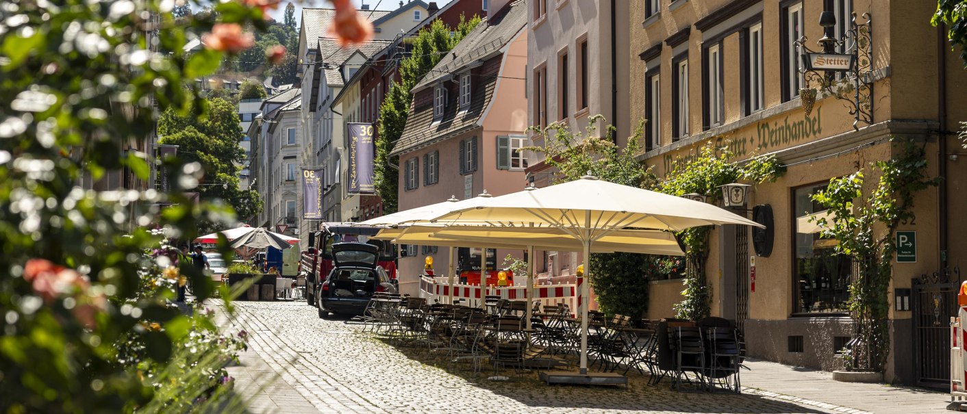 Pflasterstraße im Bohnenviertel mit Weinhaus Stetter, Außensitzbereich unter Sonnenschirmen, umgeben von blühenden Pflanzen und historischen Gebäuden., © Stuttgart Marketing GmbH, Sarah Schmid