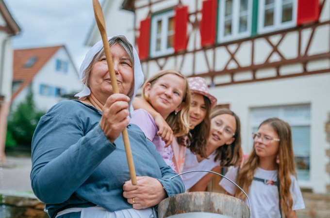Eine Frau in historischer Kleidung h&auml;lt einen Holzl&ouml;ffel, umgeben von vier Kindern. Im Hintergrund ein Fachwerkhaus mit roten Fensterl&auml;den., &copy; Stadt Waldenbuch