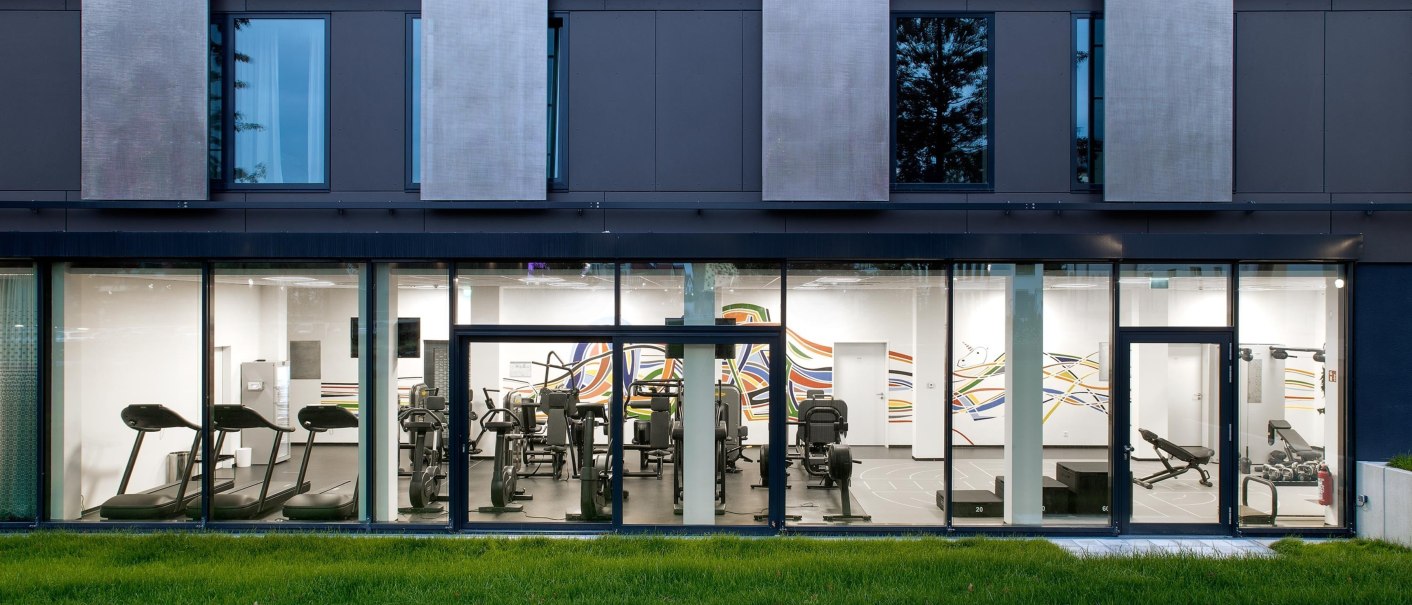 Moderne Fitnessstudio-Fassade mit großen Fenstern, die den Blick auf Laufbänder und Fitnessgeräte im Inneren freigeben. Grüne Wiese im Vordergrund., © Hotel am Remspark Moderne Fitnessstudio-Fassade mit großen Fenstern, die den Blick auf Laufbänder und Fitnessgeräte im Inneren freigeben. Grüne Wiese im Vordergrund., © Hotel am Remspark