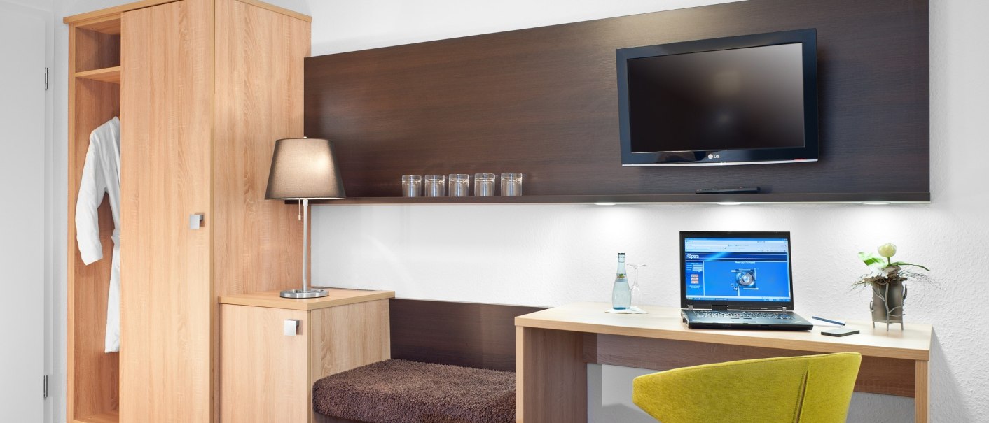 Modernes Hotelzimmer mit Schreibtisch, Laptop, gelbem Stuhl, TV, Kleiderschrank und Lampe., © TOMAS