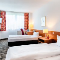 Helles Zweibettzimmer mit zwei Einzelbetten, Nachttisch mit Lampe, Schreibtisch, Fernseher und roten Vorhängen., © ACHAT Hotels Deutschland