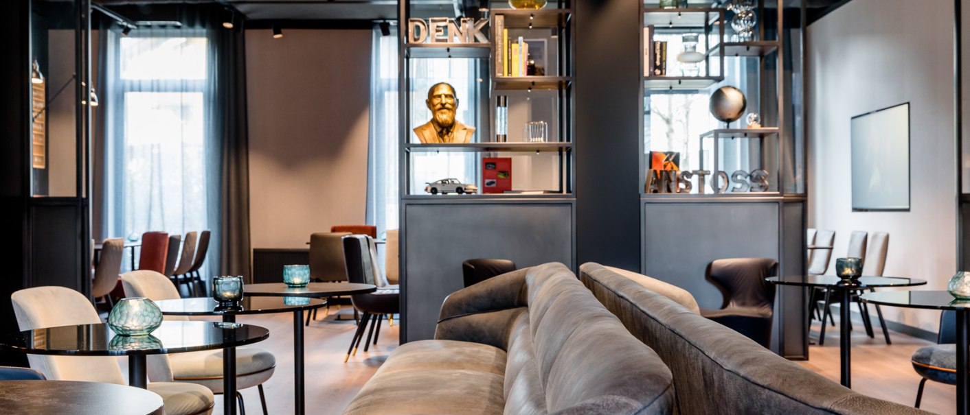 Moderne Lounge mit grauen Sofas, runden Tischen und dekorativen Elementen wie einer Büste und Büchern in Regalen., © TOMAS