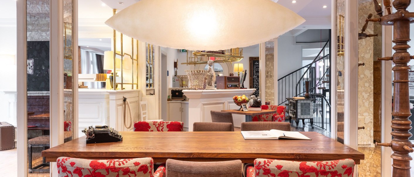 Elegante Lobby mit Holztisch, roten Stühlen und großer Deckenleuchte. Im Hintergrund eine Rezeption und eine Wendeltreppe., © Hotel Weinstadt