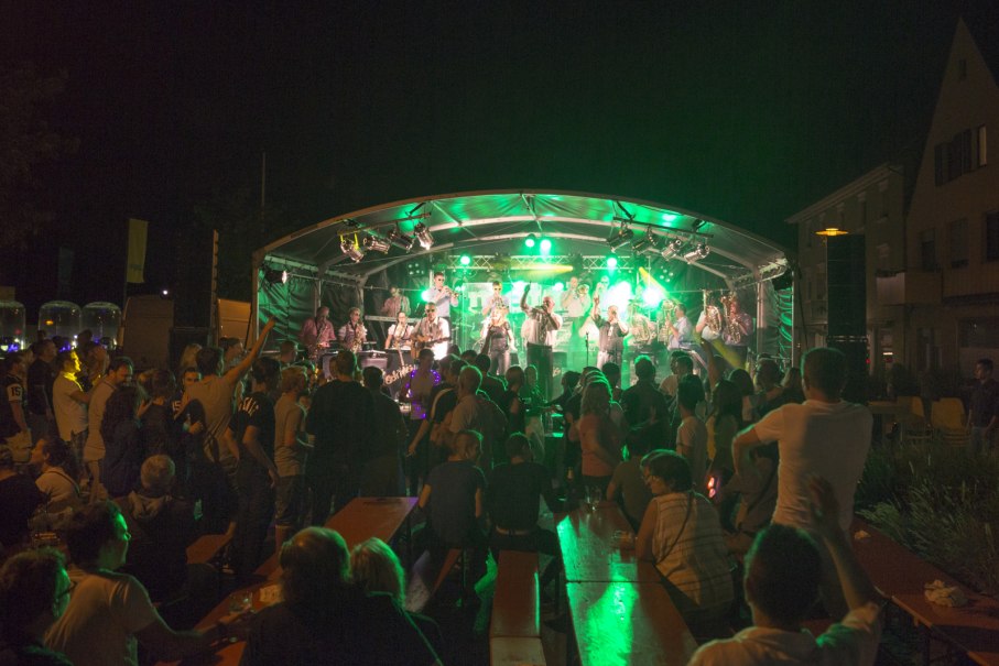 Eine Band spielt auf einer beleuchteten Bühne beim Neckarfest. Menschen tanzen und sitzen an Tischen, während sie der Musik lauschen., © D. Jüptner Eine Band spielt auf einer beleuchteten Bühne beim Neckarfest. Menschen tanzen und sitzen an Tischen, während sie der Musik lauschen., © D. Jüptner