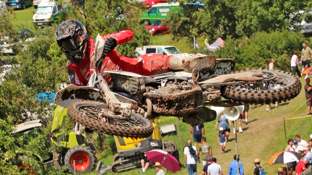 Ein Motocross-Fahrer in roter Ausr&uuml;stung fliegt mit seinem Motorrad durch die Luft. Zuschauer und Fahrzeuge sind im Hintergrund zu sehen., &copy; Rainer Kengeter