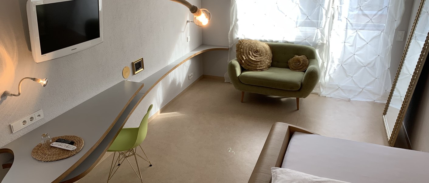 Modernes Zimmer mit grünem Sessel, Wand-TV, geschwungenem Schreibtisch und großen Fenstern mit weißen Vorhängen., © TOMAS
