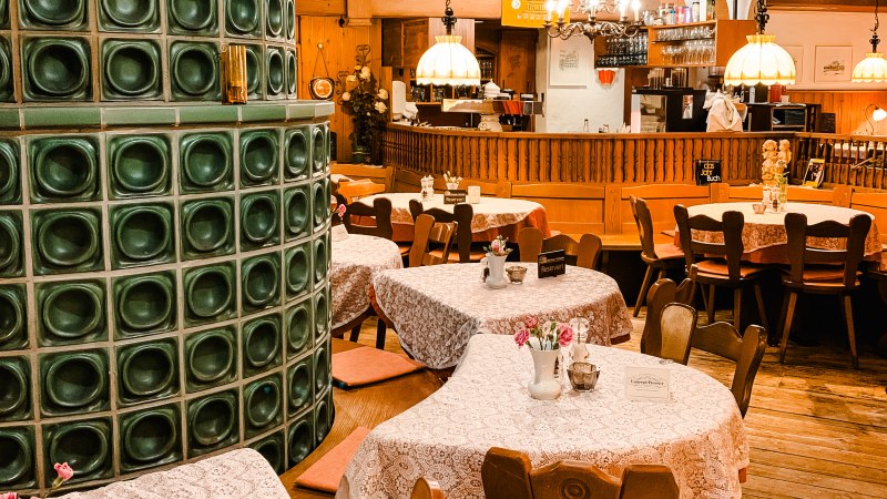 Gemütliches Restaurant mit Holzmöbeln, Tischdecken und einem grünen Kachelofen. Lampen hängen von der Decke, Bar im Hintergrund., © Weinstube Kachelofen Gemütliches Restaurant mit Holzmöbeln, Tischdecken und einem grünen Kachelofen. Lampen hängen von der Decke, Bar im Hintergrund., © Weinstube Kachelofen