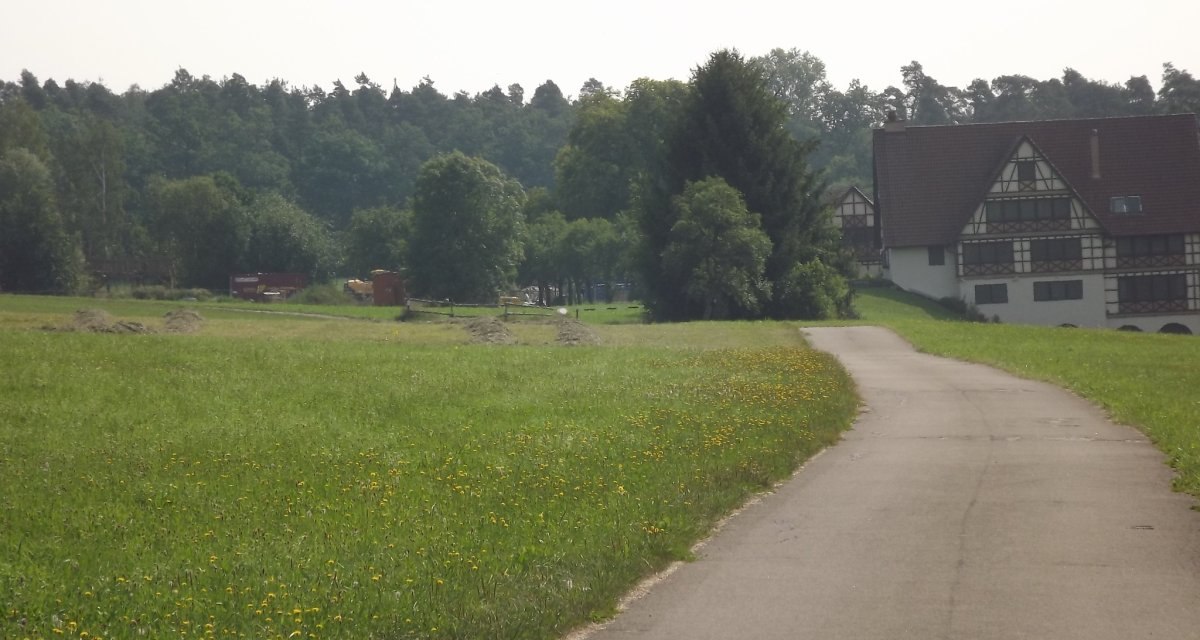 Ein ländlicher Weg schlängelt sich durch eine grüne Wiese mit gelben Blumen. Im Hintergrund steht ein großes Fachwerkhaus vor einem dichten Wald., © Natur.Nah. Schönbuch & Heckengäu Ein ländlicher Weg schlängelt sich durch eine grüne Wiese mit gelben Blumen. Im Hintergrund steht ein großes Fachwerkhaus vor einem dichten Wald., © Natur.Nah. Schönbuch & Heckengäu