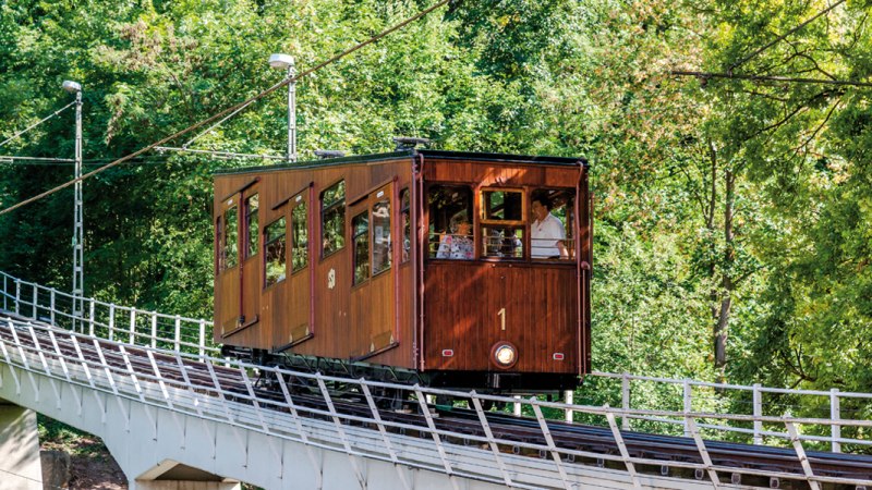 Eine historische Standseilbahn mit Holzverkleidung fährt eine geneigte Strecke entlang, umgeben von dichtem Grün., © TMBW, Gregor Lengler Eine historische Standseilbahn mit Holzverkleidung fährt eine geneigte Strecke entlang, umgeben von dichtem Grün., © TMBW, Gregor Lengler