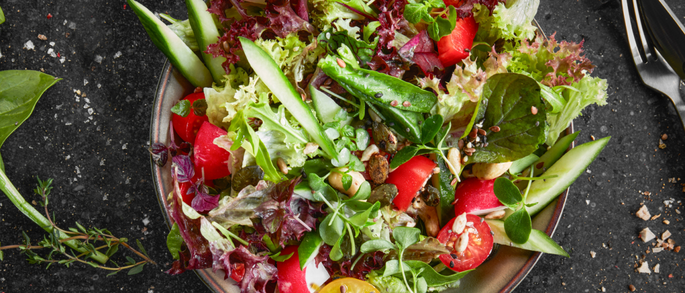 Ein bunter Salat mit Tomaten, Gurken, Blattsalaten und Kernen auf einem Teller. Daneben liegen Besteck und Kräuter auf einem dunklen Untergrund., © MAREDO