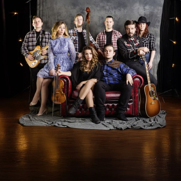 Sieben Musiker in Country-Kleidung posieren mit Instrumenten auf einem roten Sofa vor einem grauen Hintergrund., © Stadtverwaltung Schorndorf Sieben Musiker in Country-Kleidung posieren mit Instrumenten auf einem roten Sofa vor einem grauen Hintergrund., © Stadtverwaltung Schorndorf