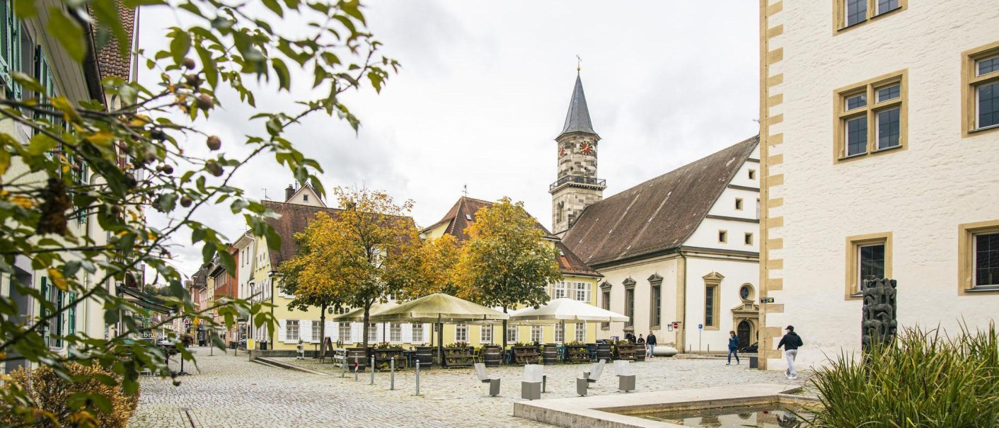 Göppinger Innenstadt mit historischen Gebäuden, einem Kirchturm und einem Platz mit Bäumen und Sitzgelegenheiten. Herbstliche Atmosphäre., © SMG, Sarah Schmid Göppinger Innenstadt mit historischen Gebäuden, einem Kirchturm und einem Platz mit Bäumen und Sitzgelegenheiten. Herbstliche Atmosphäre., © SMG, Sarah Schmid