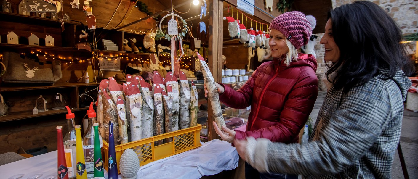 Zwei Frauen auf einem Weihnachtsmarkt betrachten handgemachte Dekorationen aus Holz an einem festlich geschmückten Stand., © Torsten Wenzler