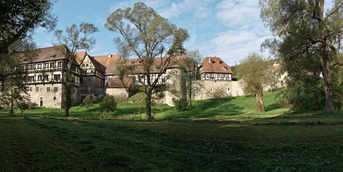 Das Bild zeigt das Kloster Bebenhausen mit Fachwerkgebäuden, umgeben von Bäumen und einer grünen Wiese unter blauem Himmel., © Günther Kleis Das Bild zeigt das Kloster Bebenhausen mit Fachwerkgebäuden, umgeben von Bäumen und einer grünen Wiese unter blauem Himmel., © Günther Kleis