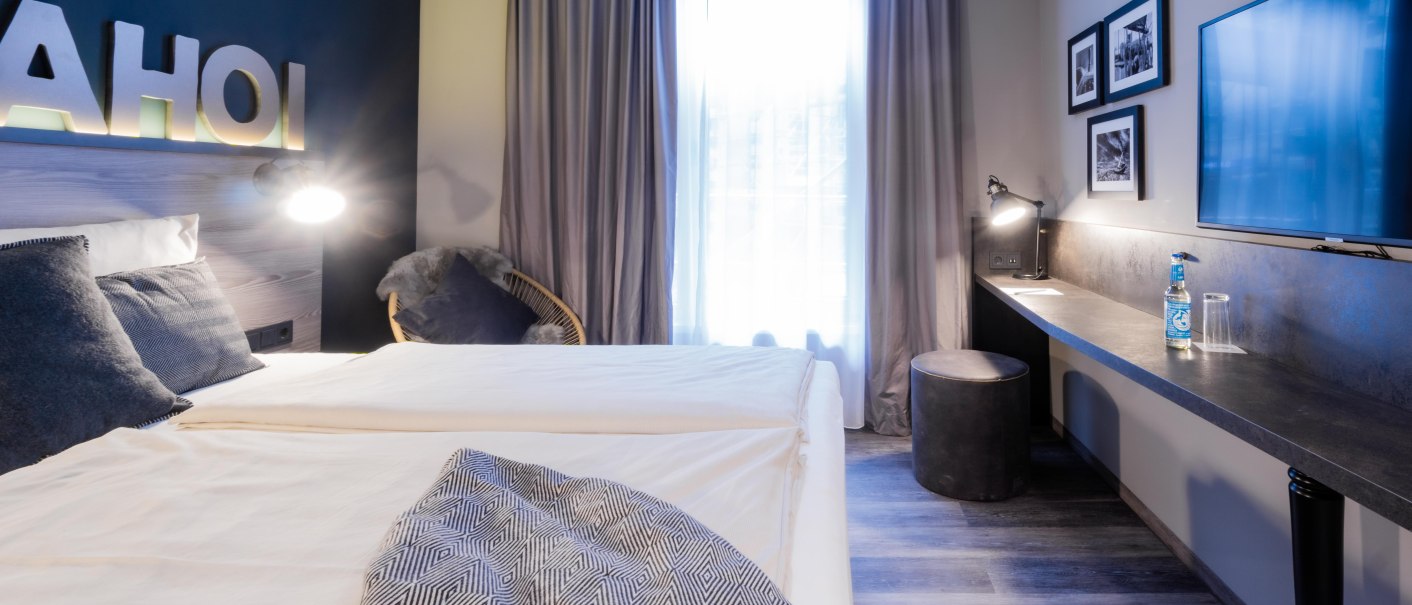 Modernes Zimmer mit Doppelbett, Schreibtisch, Flachbildfernseher und Dekoration. Schriftzug 'AHOI' an der Wand, neutrale Farben und gemütliche Atmosphäre., © HARBR. hotel Ludwigsburg / DQuadrat Living GmbH Modernes Zimmer mit Doppelbett, Schreibtisch, Flachbildfernseher und Dekoration. Schriftzug 'AHOI' an der Wand, neutrale Farben und gemütliche Atmosphäre., © HARBR. hotel Ludwigsburg / DQuadrat Living GmbH