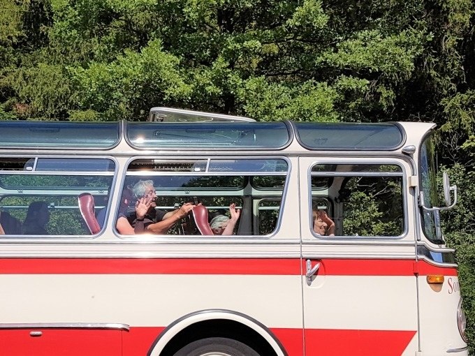 Ein Oldtimerbus mit roten und weißen Streifen fährt durch eine grüne Landschaft. Die Passagiere winken fröhlich aus den Fenstern., © SwabianTravel Ein Oldtimerbus mit roten und weißen Streifen fährt durch eine grüne Landschaft. Die Passagiere winken fröhlich aus den Fenstern., © SwabianTravel