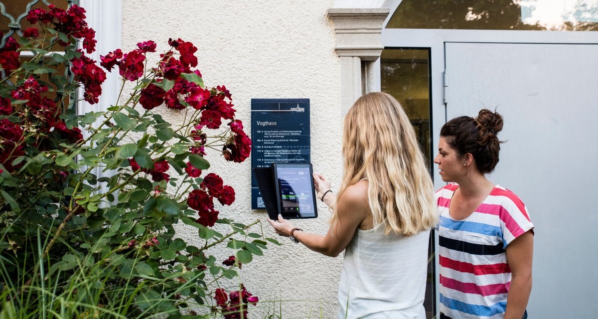Zwei Frauen stehen vor einer Tafel am Vogthaus. Eine scannt mit einem Tablet einen QR-Code. Rote Rosen bl&uuml;hen daneben., &copy; Stadt Kirchheim unter Teck