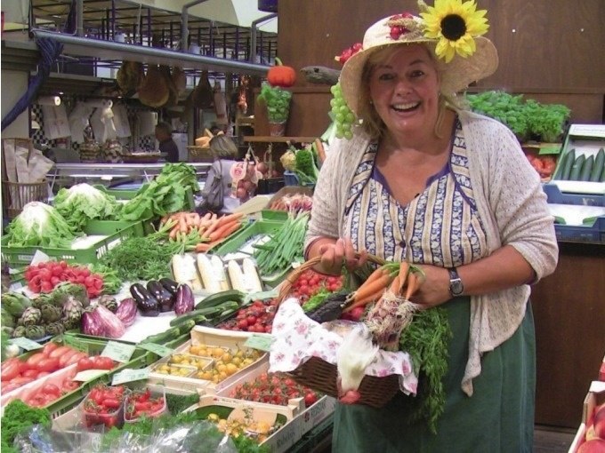 Frau mit Sonnenblumenhut in einer Markthalle, lächelnd, umgeben von frischem Gemüse und Obst. Sie hält einen Korb mit Karotten und Kräutern., © Stuttgart Marketing GmbH Frau mit Sonnenblumenhut in einer Markthalle, lächelnd, umgeben von frischem Gemüse und Obst. Sie hält einen Korb mit Karotten und Kräutern., © Stuttgart Marketing GmbH
