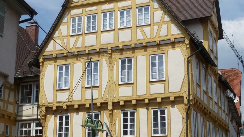 Ein historisches Fachwerkhaus mit gelben Balken, Uhr und Glockenturm. Es steht an einer gepflasterten Stra&szlig;e mit Parkscheinautomat und Blumenk&uuml;bel.