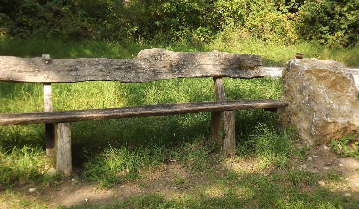 Rustikale Holzbank neben einem großen Stein, umgeben von grüner Vegetation und Gras., © Natur.Nah. Schönbuch & Heckengäu Rustikale Holzbank neben einem großen Stein, umgeben von grüner Vegetation und Gras., © Natur.Nah. Schönbuch & Heckengäu