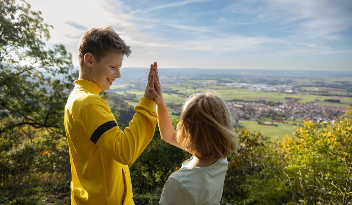 Zwei Kinder geben sich ein High-Five auf einem Hügel mit Blick auf eine weite Landschaft und einen blauen Himmel., © Foto Thomas Zehnder