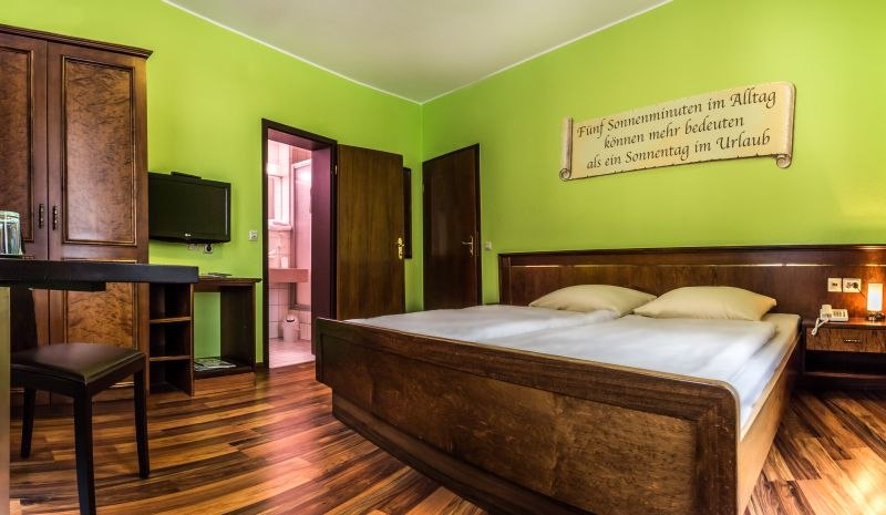 Doppelzimmer mit gr&uuml;n gestrichenen W&auml;nden, Holzbett, Schreibtisch, Fernseher und einem Spruch an der Wand. Badezimmer im Hintergrund., &copy; TOMAS