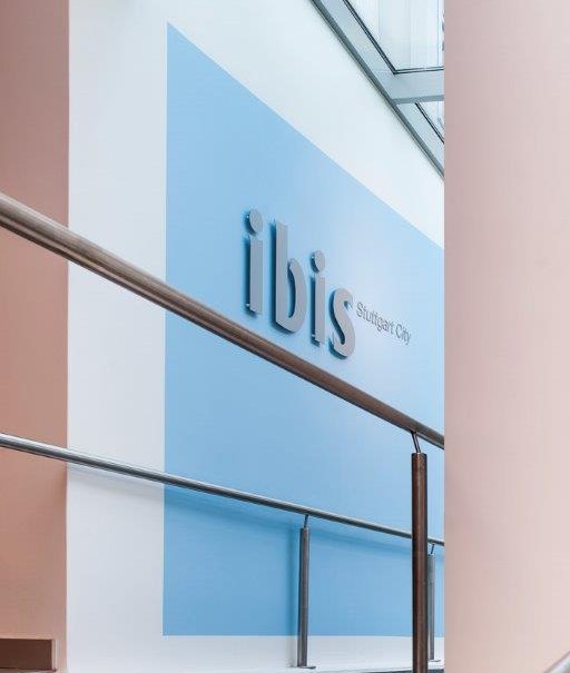 Blick auf das ibis Hotel Stuttgart City Schild in einem modernen Gebäude mit Glasdach und Handlauf., © TOMAS